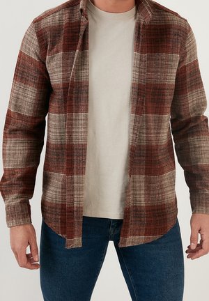 Mężczyzna w rozpiętej czerwono-beżowej koszuli w kratę na beżowym t-shircie i ciemnoniebieskich jeansach, stoi z rozluźnionymi ramionami.