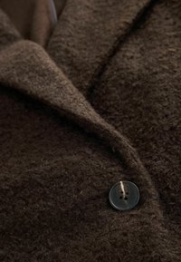 Lähikuva tummanruskeasta teksturoidusta villakoista, jossa on leveä kaulus ja yksittäinen musta nelireikäinen nappi kiinni.