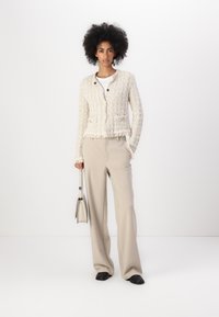 Cardigan di tessuto crema con orlo a frange e due bottoni, abbinato a pantaloni beige a gamba larga e una piccola borsa grigia. Scarpe nere completano il look.