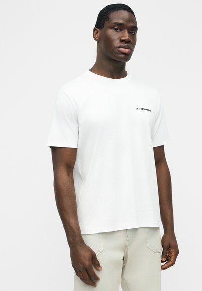 Άνδρας μοντέλο με σκούρο δέρμα που φοράει λευκό T-shirt με κοντά μανίκια και την επιγραφή "LES BENJAMINS" στο στήθος, καθώς και ανοιχτόχρωμο παντελόνι.