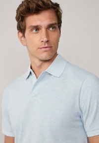 Ung man med ljusbrunt hår iförd en ljusblå kortärmad pikétröja, som tittar lätt mot höger mot en enkel bakgrund.