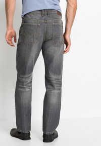 Jeans da uomo in denim scuro, con vestibilità slim, tasche posteriori con cuciture a zigzag e un leggero sbiadimento sulle cosce. Scarpe nere visibili.