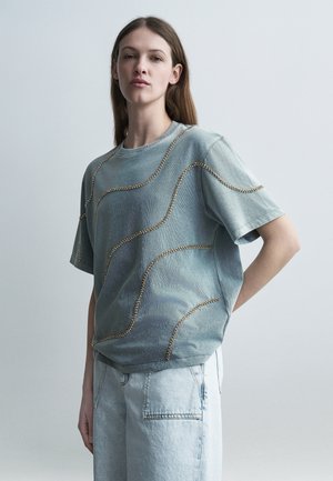 Mladá žena s dlouhými vlasy, která má na sobě volné, vybledlé modré tričko se vzorem zlatých korálkových vln a světlé džínové kalhoty.