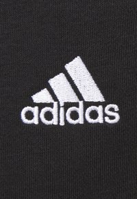 Zwart stof met een opvallend wit geborduurd Adidas-logo, met drie diagonale strepen boven het woord "adidas" in kleine letters.