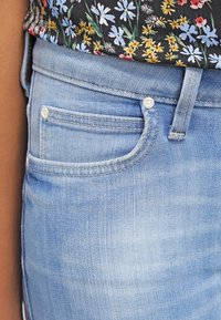 Vaqueros de mezclilla azul claro con un pequeño bolsillo delantero, sutiles desgastes en forma de rayas y detalles de costuras visibles. Tejido texturizado con una apariencia suave.