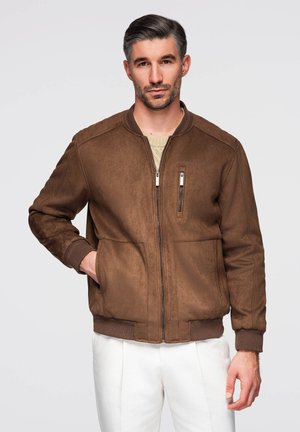 Mann mit zurückgegeltem Haar, der eine braune Wildlederjacke über einem beigen Pullover und weißen Hosen trägt, steht mit einer Hand in der Jackentasche.