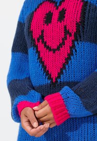 Bunt gestrickter Pullover mit einem großen pinken Smiley-Herz auf blauen Streifen. Rippbündchen in leuchtendem Pink und Marineblau mit einem groben Struktur.