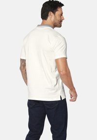 Weißes Polohemd mit blauer Kragen, kurzen Ärmeln und seitlichem Schlitz. Der Stoff wirkt glatt und eignet sich gut für legere Anlässe.