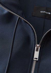 Blazer navy con una texture di tessuto liscia, dettaglio della cerniera argentata e un'etichetta nera con il nome del marchio "OPUS" vicino al colletto.