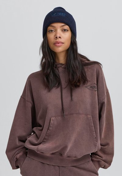 Brūns pārmērīgs hoodeis ar priekšējo kabatu, auklu un logotipa detaļu. Apvienots ar tumši zilu ribotu adītu cepuri. Vienkāršs, brīvs piegriezums.
