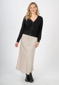 Zwarte gebreide cardigan met een V-hals en vijf knopen, gecombineerd met een crèmekleurige satijnen maxi-jurk en chunky zwarte enkellaarzen.