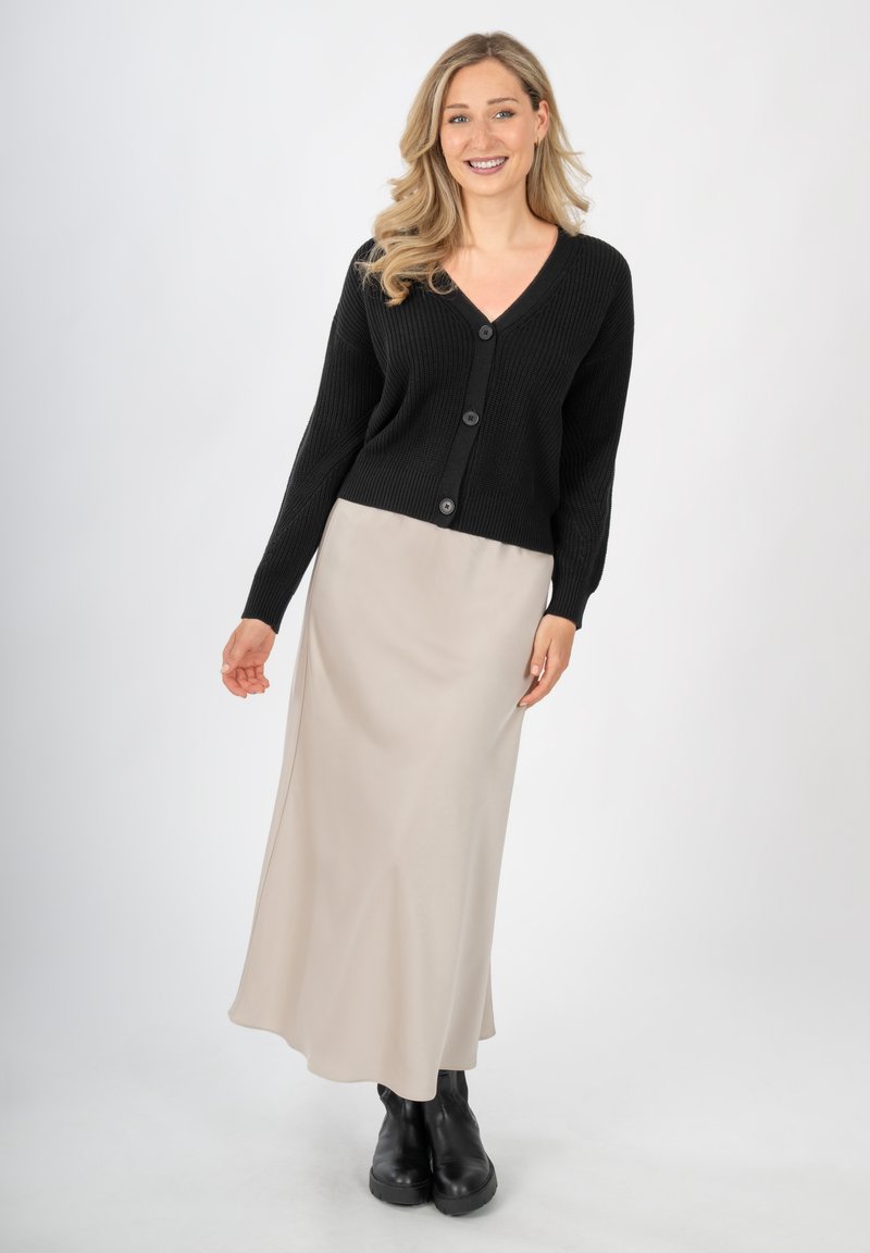 Zwarte gebreide cardigan met een V-hals en vijf knopen, gecombineerd met een crèmekleurige satijnen maxi-jurk en chunky zwarte enkellaarzen.