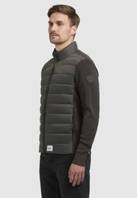 Herre tofarvet vest med en quiltet grå nylon krop og strikkede ærmer. Har en høj krave og et logo-patch på venstre arm. Lynlås lukning.