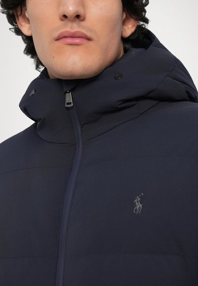 Námornícky modrá zateplená bunda s vysokým golierom a kapucňou. Obsahuje plný zips a nenápadné vyšívané logo na hrudi. Hladká textúra.