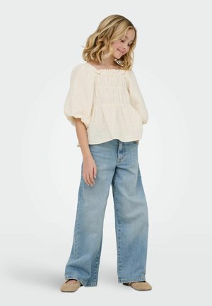 Niña joven con cabello rubio que lleva una blusa crema con mangas abullonadas, vaqueros anchos de color azul claro y zapatos planos beige, de pie contra un fondo liso.