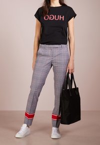 Maglietta nera con logo rosso "HUGO", pantaloni a quadri grigi con striscia rossa, sneakers bianche e una borsa tote nera. Texture lisce e piatte.