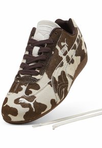 Ténis com estampado de vaca castanho e branco, com uma textura suave, apresentando atacadores escuros, marcação em destaque e uma sola de borracha de perfil baixo.