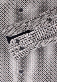 Overhemd met knopen, voorzien van een geometrisch patroon in zwart, bruin en wit. Close-up van de manchette met zwarte knopen, die de textuur van de stof toont.