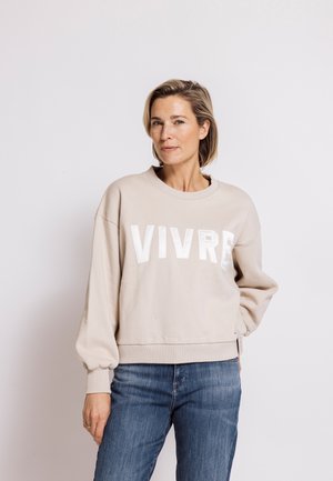 Frau mit kurzen blonden Haaren, die einen beigen Pullover mit dem Text "VIVRE" und blaue Jeans trägt, steht mit der Hand in der Tasche vor weißem Hintergrund.