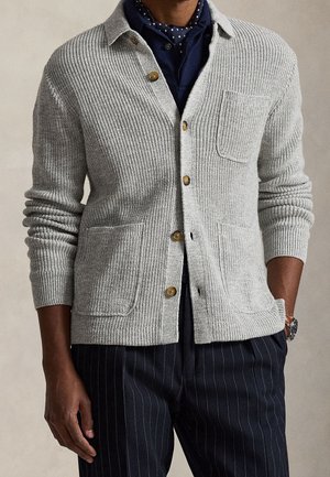 Man wearing a light gray buttoned knit cardigan with three front pockets, over a dark shirt with a patterned scarf and pinstriped pants.