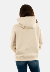 Sudadera beige con una gran capucha ajustable, textura suave y puños acanalados, combinada con jeans de color azul oscuro. Detalles de costura visibles en la parte posterior.