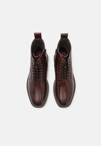Une paire de bottines en cuir verni marron foncé à lacets, avec fermeture éclair sur le côté et languettes de tirage, sur un fond blanc.