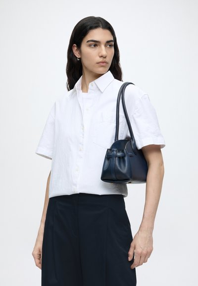 Femme aux longs cheveux bruns porte une chemise blanche à manches courtes boutonnée et un pantalon noir, portant un petit sac à main en cuir bleu marine sur son épaule.