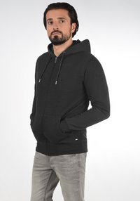 Sort zip-up hoodie lavet af tekstureret stof, med hætte, snore og frontlommer. Bæret med lysegrå jeans.