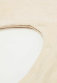 Weiches beiges Kleidungsstück mit einem runden Ausschnitt und Druckknopf-Verschlüssen. Der Stoff wirkt strukturiert, mit einer glatten Oberfläche an den Kanten.