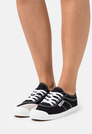 Jambes portant des baskets en toile noires et blanches à lacets avec des semelles blanches sur un fond clair uni.