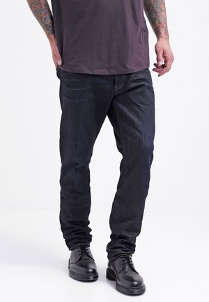 Personne portant un jean bleu foncé, des chaussures en cuir noir à lacets, et un t-shirt violet foncé, debout les mains détendues le long du corps.