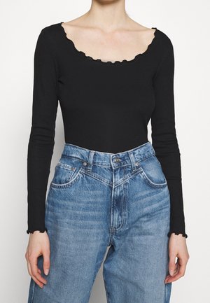 Žena oblečená v černém topu s dlouhými rukávy s ozdobným vroubkovaným výstřihem a manžetami, spojeném s modrými džíny s vysokým pasem.