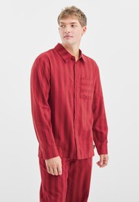 Camisa de manga larga a rayas rojas con cuello puntiagudo y un bolsillo en el pecho. Textura suave, cierre de botones al frente y corte relajado.