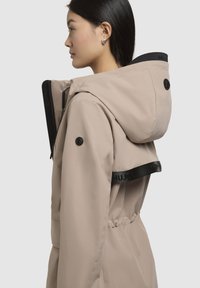 Beige vandtæt jakke med hætte, prydet med sorte detaljer og hardware, sidelukning med lynlås og en indsnøret talje for et tætsiddende udseende.