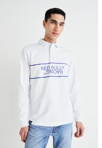 Balta garām piedurknēm polo krekls ar apkakli un zilu "RED BULL RACING" grafiku uz krūšu; valkāts ar gaiši ziliem džinsiem.