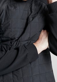 Svart kviltad jacka med en texturerad yta; har långa ärmar med ribbade muddar och dragkedja.