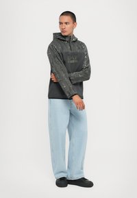 Hættetrøje i mørkegrå med strukturerede ærmer, med et sort frontpanel og grønt logo. Sammen med lyseblå brede jeans.