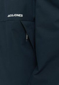 Sötétkék dzseki sima textúrával. Oldalsó zsebbel rendelkezik, és a mellkason fehér "Jack & Jones" logó található, amely kiemeli a márkát.