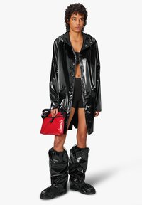 Abrigo de lluvia negro y brillante con cierre de botones, acompañado de botas negras impermeables. Sosteniendo un bolso pequeño rojo con una correa negra.