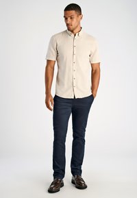 Man in een beige kortemouw overhemd, donkere blauwe broek en bruine leren schoenen, staand met een hand in zijn zak en kijkt naar beneden.