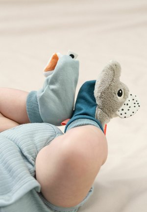 Chaussons plush bleus et gris avec des caractéristiques animales, comprenant un nez texturé et des oreilles, portés aux pieds d'un enfant sur un fond clair.