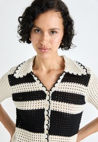 Hæklet cardigan med skiftende sorte og creme striber, bølget krave og træknapper. Korte ærmer og struktureret strikdesign.