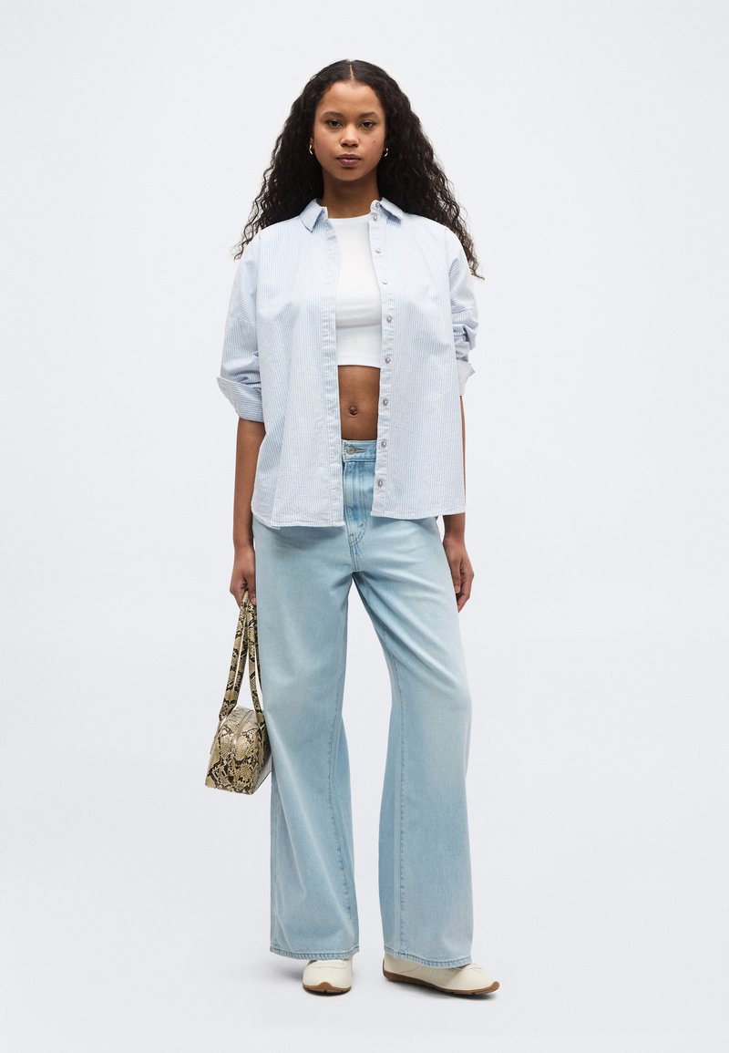 Femme aux cheveux longs et bouclés portant une chemise à rayures bleu clair par-dessus un crop top blanc, un jean large bleu clair, des chaussures blanches et tenant un sac à main en cuir de serpent.
