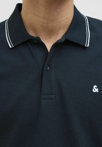 Tumedat meresärgid tekstuuriga kangast, millel on kahe nupuga kaelus, valged triibud kaeluses ja väike valge tikandiga ampersand logo.