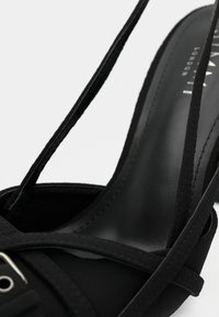 Čierna sandála na vysokom opätku s prekríženými remienkami, hladkým matným povrchom a elegantnou špicatou špičkou; obsahuje jemný kovový detail na pracke.