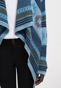 Cardigan en maille à motifs bleus avec des dessins géométriques et rayés, porté sur un haut blanc et un pantalon noir, montrant la taille et le bras droit.