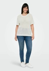 Hvid strikket top med et tekstureret diamantmønster, kortærmede, og bølget kant, parret med blå jeans og hvide sneakers.