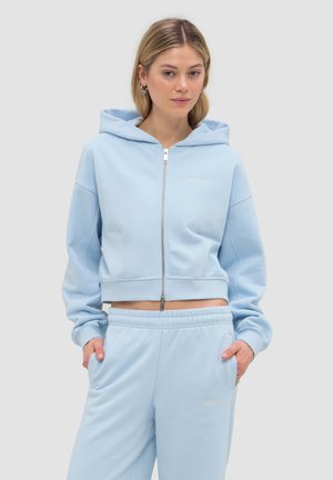 Junge Frau trägt hellblauen Kapuzenpullover mit Reißverschluss und passende Jogginghose, mit den Händen in den Taschen, steht vor einem schlichten Hintergrund.