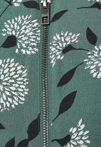 Grön zip-up-plagg med en texturerad tyg och ett blommönster i vitt och svart, centrerat runt en silverdragkedja.