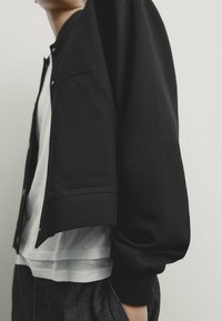 Personne portant une veste noire boutonnée par-dessus une chemise blanche et un jean foncé, debout devant un fond clair uni.